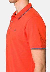 Koszulka polo z krótkim rękawem w jasnopomarańczowym kolorze z teksturowanej tkaniny. Posiada dwie guziki przy dekolcie oraz kontrastowe niebieskie wykończenie kołnierzyka i rękawów.
