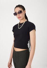 Black fitted cotton T-shirt with short sleeves, paired with olive green ribbed pants. Accessories include layered silver necklaces and oversized sunglasses.