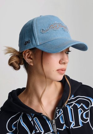 Joven con múltiples perforaciones en las orejas y anillo en la nariz lleva una gorra Ed Hardy azul claro y sudadera negra con diseño gráfico azul y blanco.