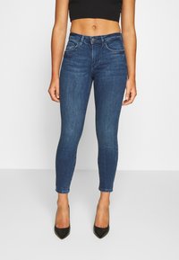 Dark blue skinny jeans with a high waist, five pockets, and visible stitching details, paired with black pointed heels.