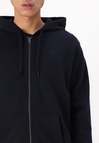 Mornarica zip-up kapuca izdelana iz mešanice bombaža, z kenguru žepom, vrvico in diskretnim logotipom na prsih.