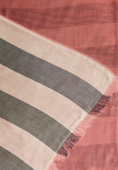 Απαλό ύφασμα με φαρδιές οριζόντιες ρίγες σε μπεζ και σκούρο γκρι, απλωμένο πάνω σε απαλό ροζ φόντο με υφή και φράντζες στο περίγραμμα.