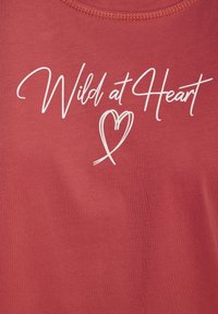 Červené bavlnené tričko s bielym písaným nápisom "Divoký v srdci" a srdcovou grafikou, okrúhlym výstrihom a krátkymi rukávmi.