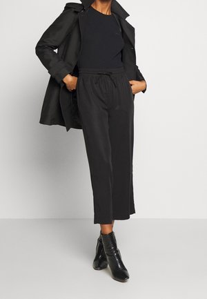 Frau trägt ein schwarzes Langarmshirt, schwarze cropped Pants mit Kordelzug, einen schwarzen Trenchcoat und schwarze Ankle Boots mit strukturiertem Muster.