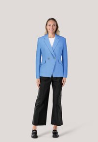 Helles blaues, doppelt geknöpftes Sakko aus strukturiertem Stoff mit zwei vorderen Taschen und silbernen Knopfakzenten, kombiniert mit schwarzen weit geschnittenen Hosen.
