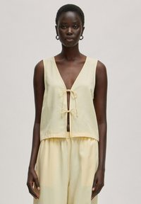 Lichtgele mouwloze top met een diepe V-hals en striksluiting aan de voorkant. Zacht materiaal met een losse pasvorm en subtiele textuur.