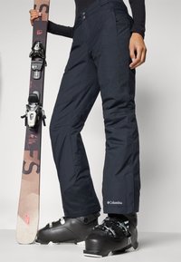 Sorte isolerte snøbukser med en glatt tekstur, med to knapper, sidelommer og Columbia-logo ved faldet; kombinert med skistøvler og en ski.