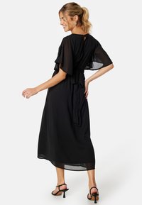 Schwarzes, bodenlanges Kleid aus leichtem Stoff. Verfügt über flatternde Ärmel, einen Schlüsselloch-Rücken und einen Bindegürtel in der Taille. Glatte Textur.