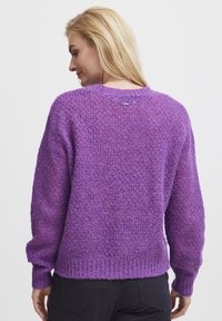 Pull en tricot violet avec une finition texturée, ourlet et poignet côtelés, comprenant un détail d'étiquette au dos. Manches longues, coupe décontractée.