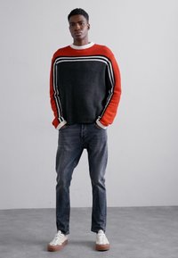 Maglione a righe rosse e nere in maglia con maniche lunghe, indossato con jeans blu e sneakers bianche con suole marroni, in piedi davanti a uno sfondo chiaro.