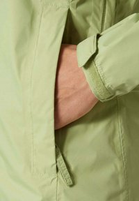 Jasnozielona kurtka o miękkiej fakturze, z boczną kieszenią i regulowanym mankietem. Widoczne ręce spoczywające w kieszeni.