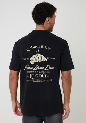 Mężczyzna w czarnej koszulce z beżową grafiką rogalika i napisem francuskiej piekarni na plecach, stojący na tle jasnego, jednolitego tła.