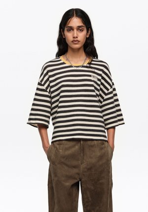 Mujer con cabello largo y oscuro, vistiendo un suéter suelto de rayas negras y blancas y pantalones marrones de cintura alta, con las manos en los bolsillos, fondo neutro.