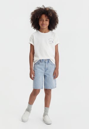 Enfant aux cheveux bouclés portant un t-shirt blanc, un short en jean clair, des chaussettes grises et des baskets blanches sans lacets, debout devant un fond uni.