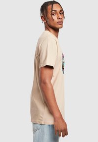 Beige bomull T-skjorte, kortermet, avslappet passform. Har et fargerikt grafisk trykk på fronten. Brukt sammen med lyseblå denimjeans.