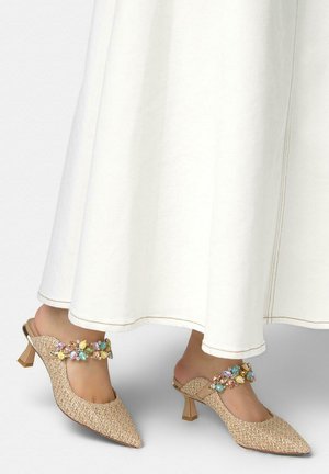 Mules tejidos beige con punta en pico y tacón, adornados con piedras de colores, usados con una falda blanca hasta el tobillo.