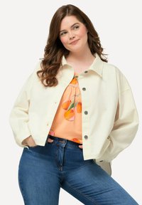 Crème-kleurige oversized jas met een kraag, knoopsluiting en textuurstof, gedragen over een perzikkleurig top met fruitprint en blauwe jeans.
