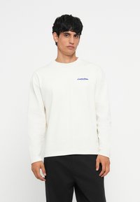 T-shirt crème à manches longues avec un col rond, mettant en avant un logo bleu brodé sur la poitrine gauche ; associé à un pantalon noir.