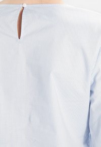 Chemise rayée bleu clair et blanche avec un col rond, une fermeture par boutons au dos et une petite découpe en forme de goutte à l\'arrière supérieur. Texture douce.