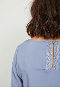 Ljusblå blus med rundad halsringning, spetsdetaljer längs ryggen och en knappknäppning högst upp vid en liten nyckelhålöppning.