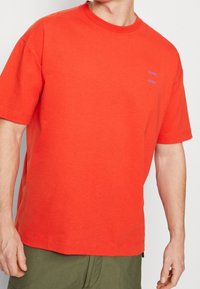 T-shirt en coton rouge avec un col rond, des manches courtes et une coupe décontractée. Présente un logo bleu discret sur la poitrine. Tissu texturé.