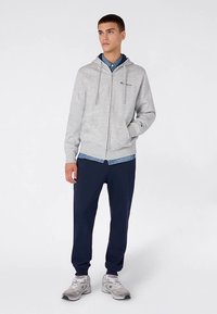 Grijze hoodie met rits en trekkoord, marineblauwe joggingbroek en zilveren sneakers. Gelaagd met een blauw overhemd met knopen eronder.