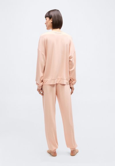 Γυναίκα με κοντά σκούρα μαλλιά, φοράει φαρδιά ρόμπα μακρυμάνικη σε χρώμα ροδάκινο και ταιριαστό παντελόνι με φαρδιά πόδια, στέκεται ξυπόλυτη γυρισμένη προς τα πίσω.