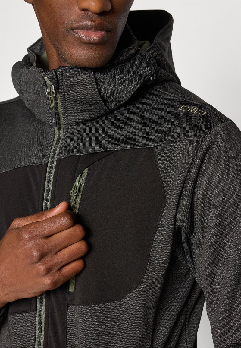 Giacca a zip grigia e nera con tessuto texture, colletto alto e tasca laterale, caratterizzata da dettagli in verde sulle cerniere e branding.