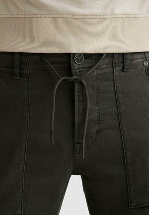 Close-up van een zwarte broek met een knoop, trekkoord en zijzak met patch, gedragen met een beige geribbelde top met een tailleband.