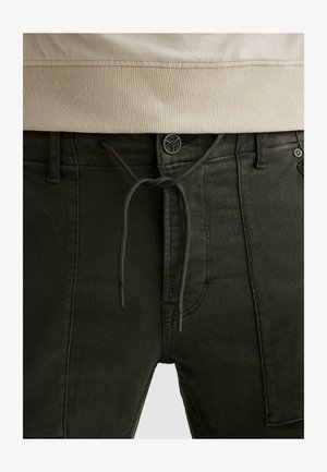 Close-up van een zwarte broek met een knoop, trekkoord en zijzak met patch, gedragen met een beige geribbelde top met een tailleband.