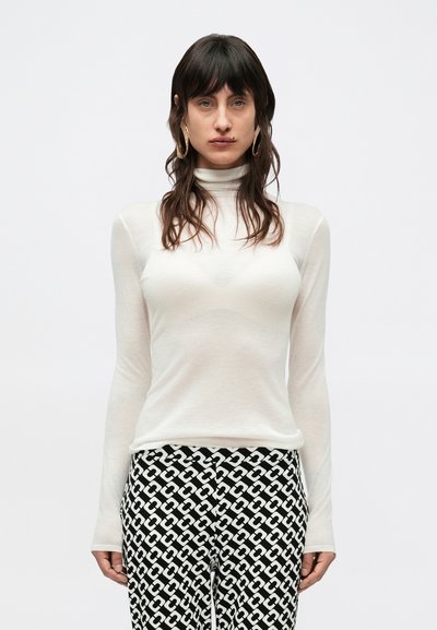 Haut blanc à manches longues et col roulé en matériau transparent, associé à un pantalon noir et blanc à motifs géométriques.