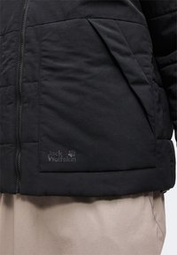 Veste matelassée noire avec fermeture éclair verticale et grande poche avant, affichant un logo Jack Wolfskin discret au-dessus d’un pantalon beige.