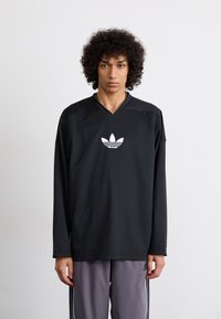 Mustapaita, jossa on pitkät hihat ja V-pääntie, ja jossa on valkoinen adidas-logo rinnassa. Valmistettu teksturoidusta kankaasta, yhdistetty harmaisiin housuihin.