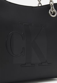 Sac à main noir en cuir lisse, avec un logo "CK" en relief, des accents en chaîne argentée et un design structuré avec un bord supérieur arrondi.