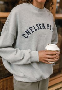 Šedá mikina se vyšitým nápisem "CHELSEA PE" v tmavě modré barvě, volný střih, žebrované manžety, nošená s jednorázovým kelímkem s kávou v ruce.