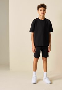Conjunto de camiseta y shorts de algodón negro, con un ajuste relajado y mangas cortas, combinado con zapatos deportivos blancos y calcetines blancos acanalados.