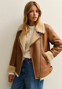 Giacca di pelle marrone con colletto e polsini in shearling crema, dotata di chiusura con zip e tasche angolate, indossata sopra un maglione in cotone crema.