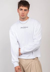 Valge pika varrukaga t-särk ümmarguse kaelusega, mille rinnal on peen must trükitäht. Pehmest kangast, vaba lõikega, lihtsa disainiga.