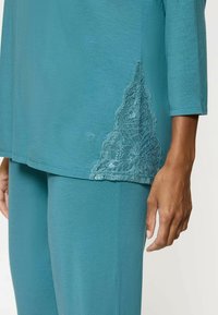 Tunika teal con maniche a ¾, caratterizzata da un delicato dettaglio in pizzo sul lato sinistro inferiore. Tessuto morbido e vestibilità comoda.