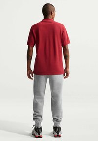 Bărbat cu tatuaje pe brațe, purtând un tricou roșu cu mâneci scurte, pantaloni de trening gri deschis și adidași negri cu alb, stând cu fața spre spate.