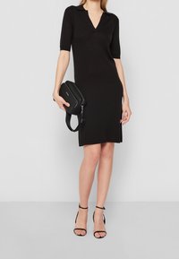 Robe noire en tricot avec un col en V et des manches courtes, offrant une coupe ajustée et des fentes latérales, assortie à des talons noirs à brides et un petit sac.