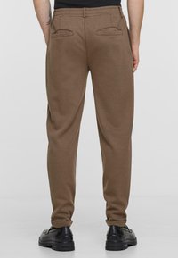 Brown, stretchy jogger pants with a soft texture, elastic waistband, and two back pockets. Worn with black shoes and rolled cuffs.