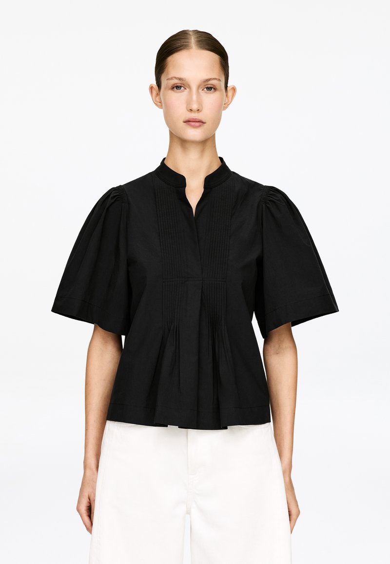 Blouse en coton noir avec col montant, manches courtes bouffantes et devant plissé, présentant une coupe décontractée et une texture lisse.