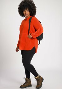 Fleecepullover i lys orange med en forlommeklappe og sideslids, parret med sorte leggings og brune vandrestøvler. Sort rygsæk bæres.