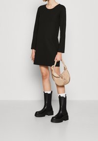 Svart långärmad klänning, slätt tyg, rund halsringning. Modellen håller en beige kuvertväska. Bär kraftiga svarta ankelstövlar med vita strumpor.