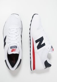 Weiße Sportschuhe mit einem Mesh-Obermaterial, schwarzen Akzenten und roten Details an der Sohle. Mit einem auffälligen "N"-Logo und gepolstertem Kragen.