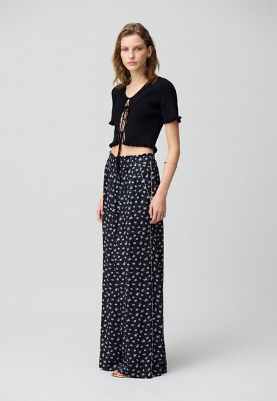 Suéter negro corto con mangas cortas acampanadas, combinado con una falda larga negra de cintura alta con estampado floral que presenta patrones en blanco y una abertura lateral.
