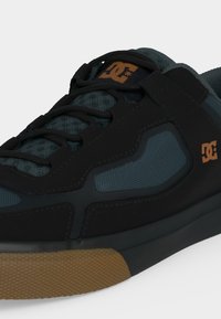 Gros plan sur une basket DC noire et vert foncé avec un logo marron et une semelle en caoutchouc marron texturée.