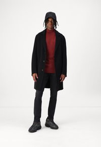 Sort frakke med knapper, rød struktureret sweater, mørke jeans og sorte sneakers med lagdelt design og strukturerede såler. Sort kasket med logo.