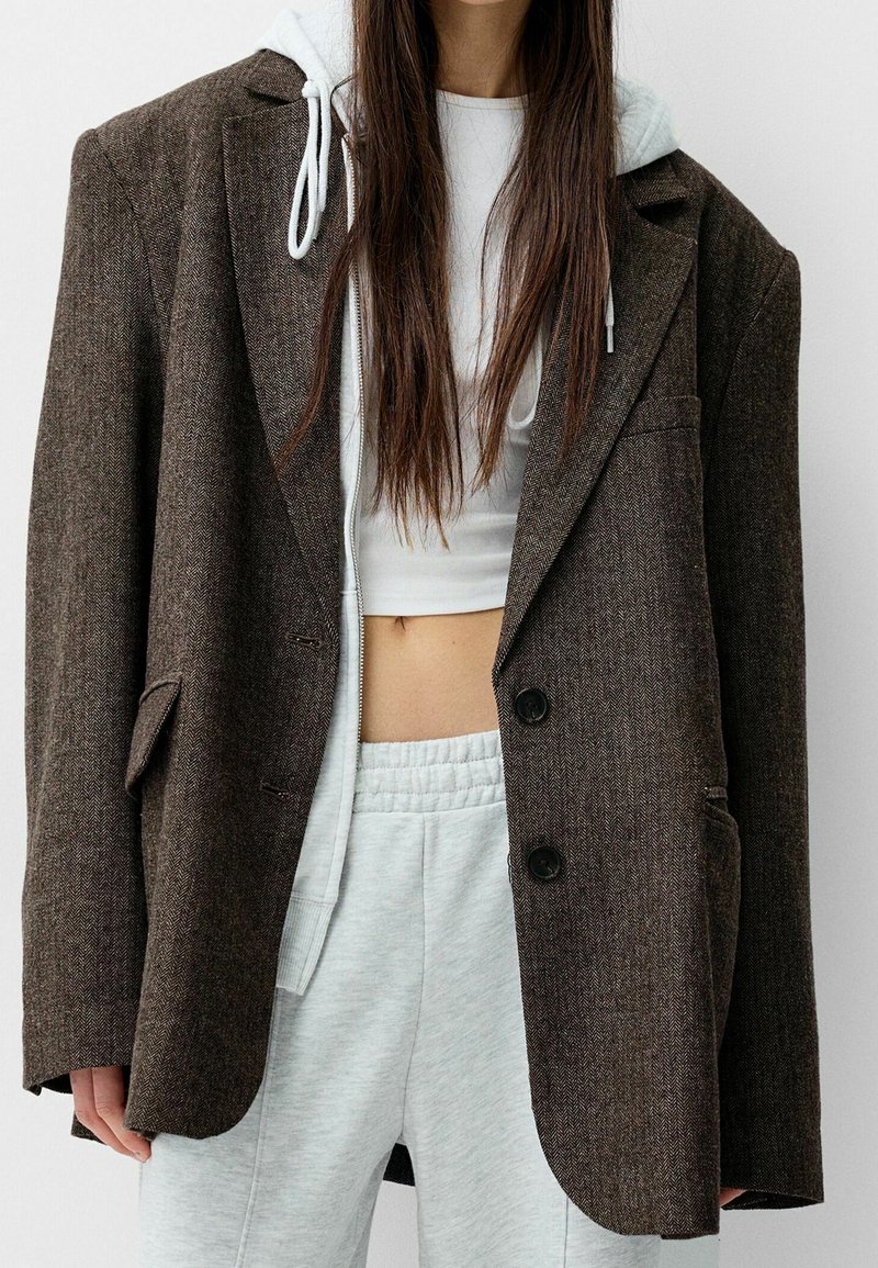 Blazer a motivi herringbone marrone con rever a pistagna, chiusura a un bottone e tasche frontali, indossato sopra una maglietta corta bianca e pantaloni della tuta grigi.